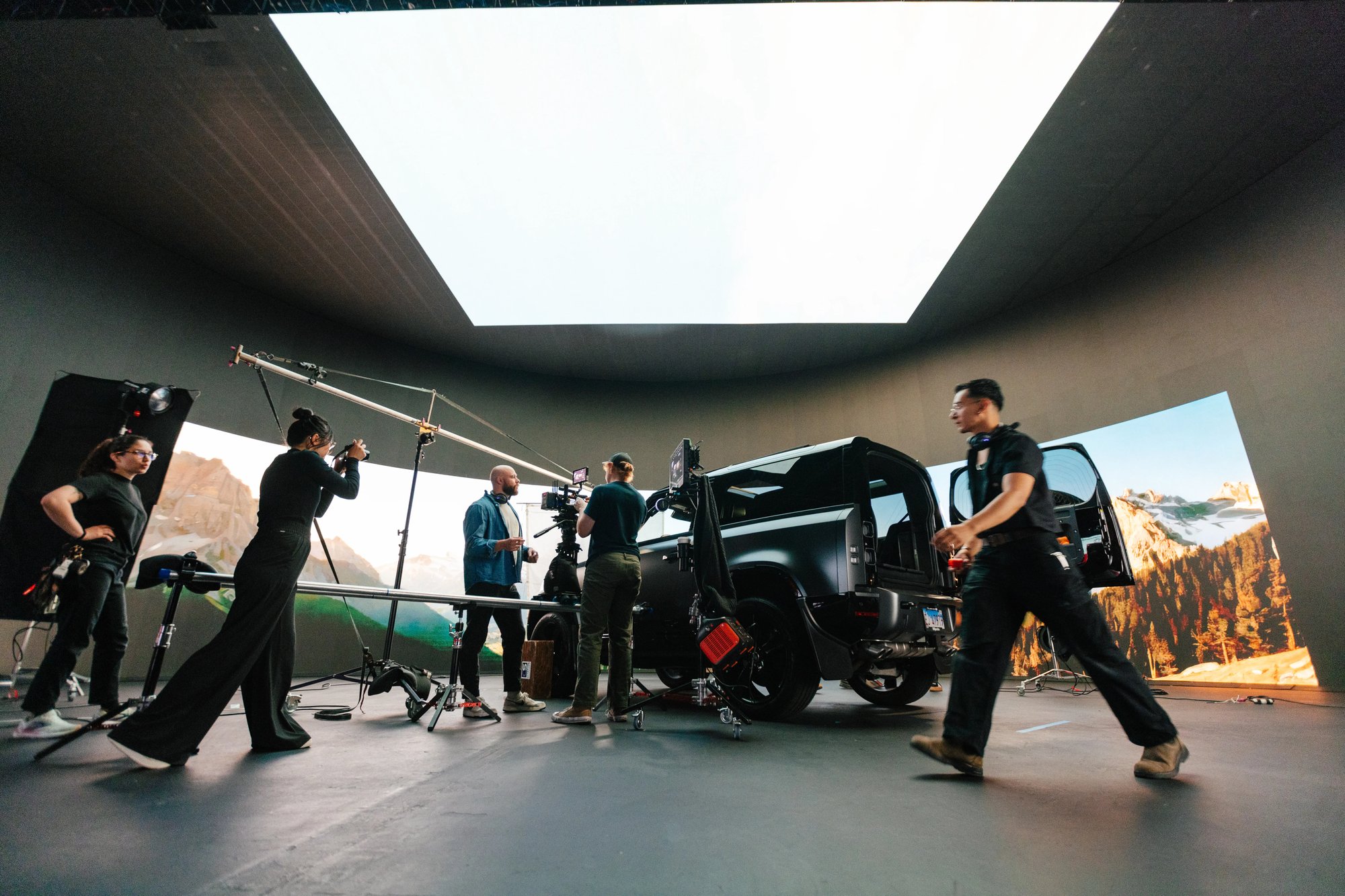 Film crew working in a studio filming a vehicle scene, with lighting rigs, cameras, and a large overhead light panel illuminating a mountain backdrop.