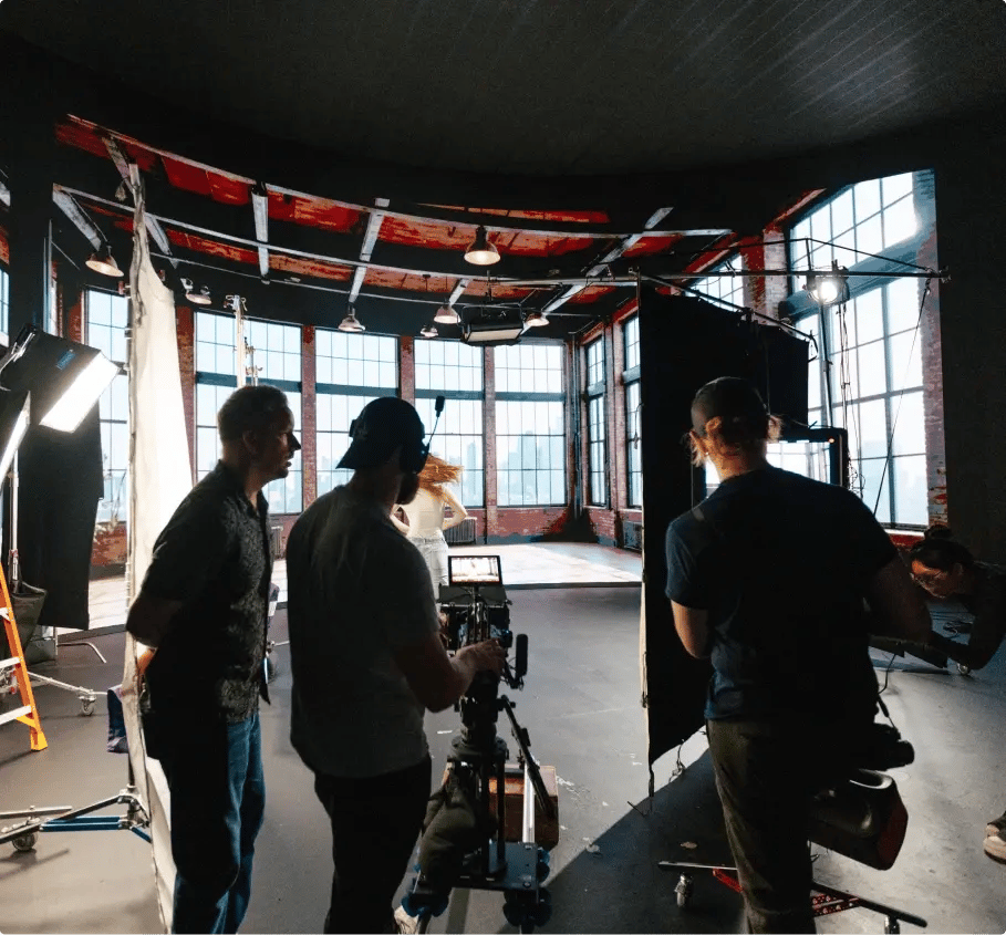 Film crew setting up cameras and lighting equipment inside a large studio with tall windows during a video production shoot.