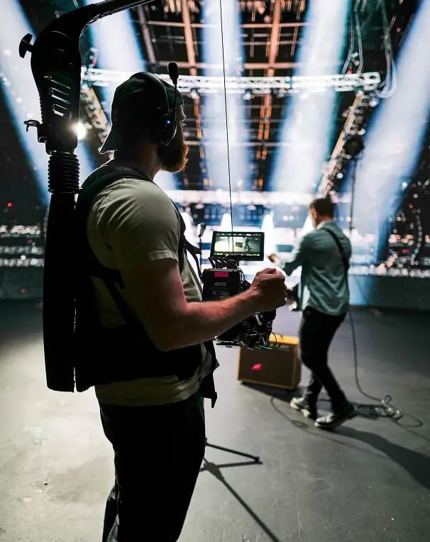 Camera operator wearing a stabilizer rig films a performer on stage under dramatic concert lighting beams.