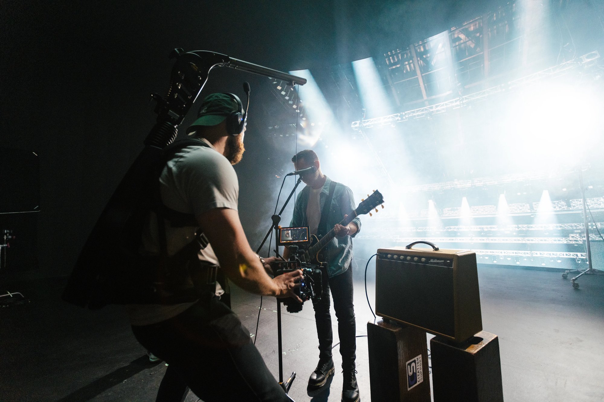Behind-the-scenes shot of a cameraman filming a guitarist performing on stage under bright concert lighting, with an amplifier and microphone visible.
