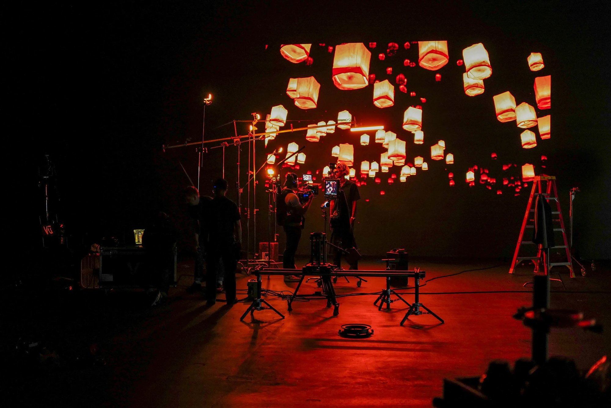 Behind-the-scenes film set with a crew preparing a scene under glowing red lantern lights, surrounded by camera equipment and rigging.